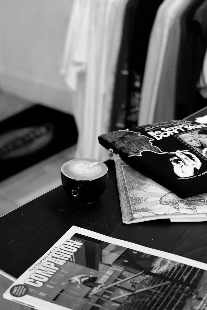 A cup of coffee with a heart-shaped latte art pattern sits beside some magazines.